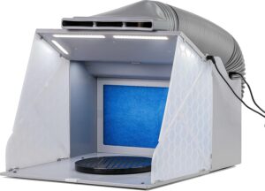 The image shows a portable airbrush spray booth with a bright blue filter, rotating turntable, and overhead LED lights. An attached exhaust hose directs fumes away, making it ideal for indoor painting projects that require good ventilation and precision.