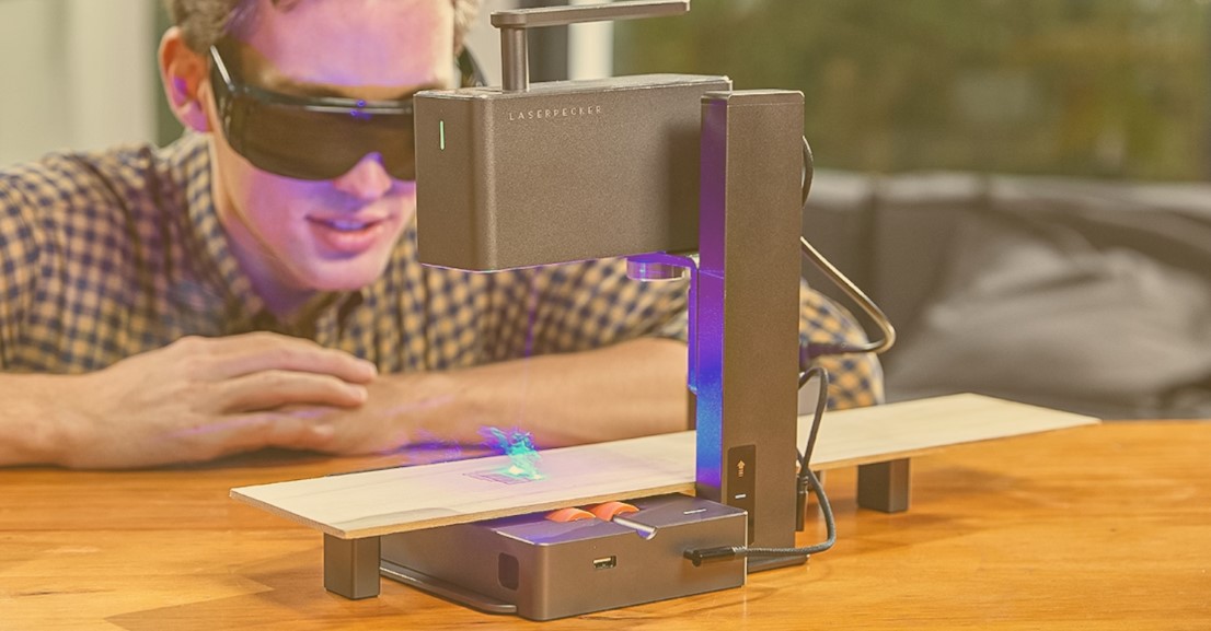 A portable laser engraver in action, engraving a piece of wood on a wooden table with a person observing in the background.