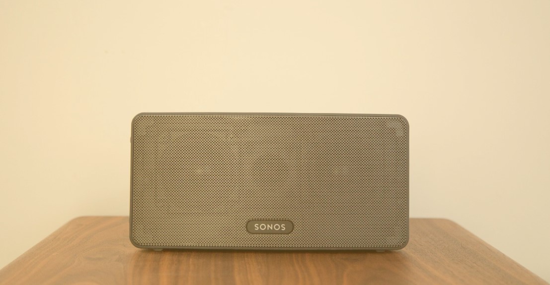 A Sonos speaker placed on a wooden surface against a plain beige background. The speaker is rectangular with rounded edges and has a mesh front with the Sonos logo in the center.