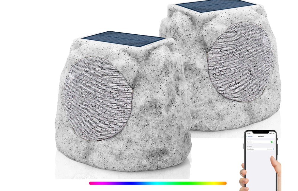 Two rock-shaped outdoor speakers with solar panels on top. The speakers have a stone-like appearance and a mesh covering the speaker area. A smartphone in the image displays Bluetooth settings, indicating wireless connectivity. A color gradient bar at the bottom suggests LED lighting features.