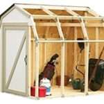 A partially constructed wooden shed with an open front, showing various gardening tools and equipment inside, including a wheelbarrow, shovel, rake, golf bag, and cooler. The shed has a slanted roof and wooden beam frame, with the front wall and door still to be installed.