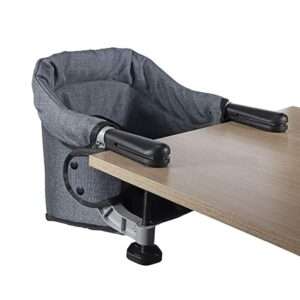 A portable restaurant high chair attached to a wooden table. The high chair is made of grey fabric with black armrests and a sturdy clamp mechanism for secure attachment.